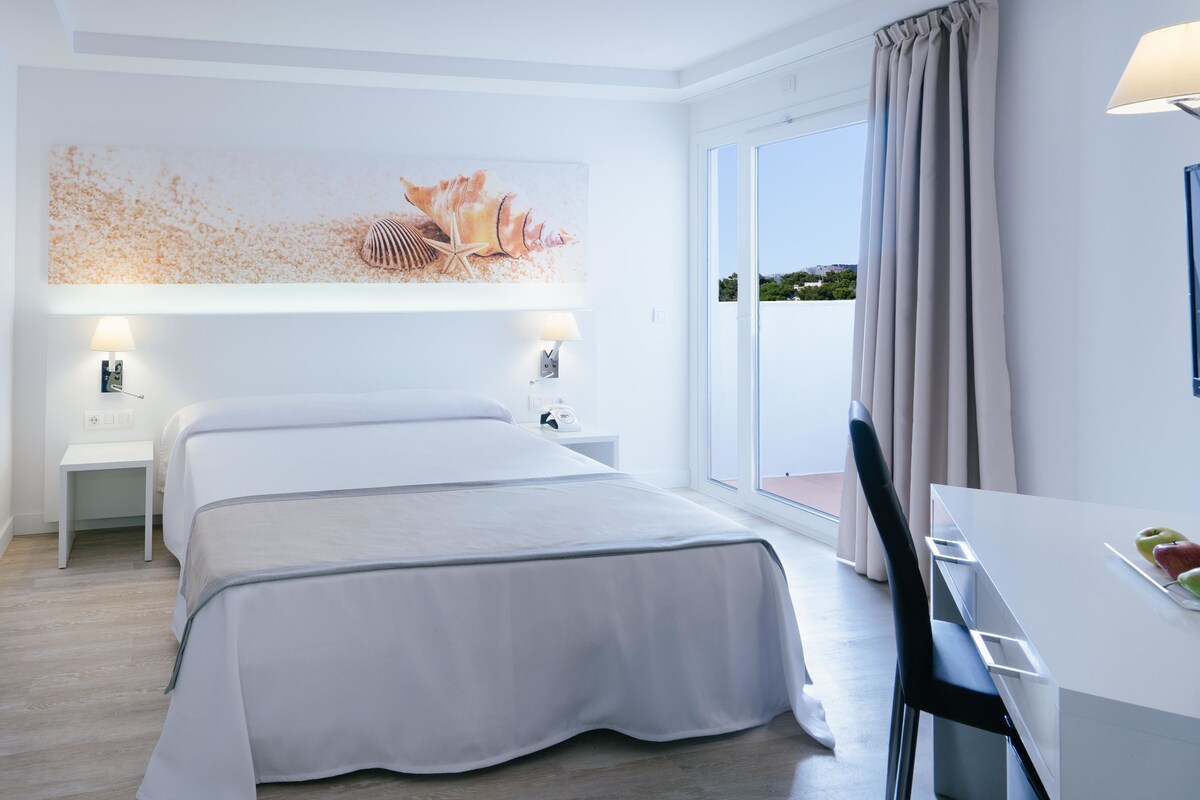 Ein modernes Hotelzimmer mit einem großen Bett, einem Schreibtisch mit Stuhl und Blick auf die Stadt durch das Fenster.