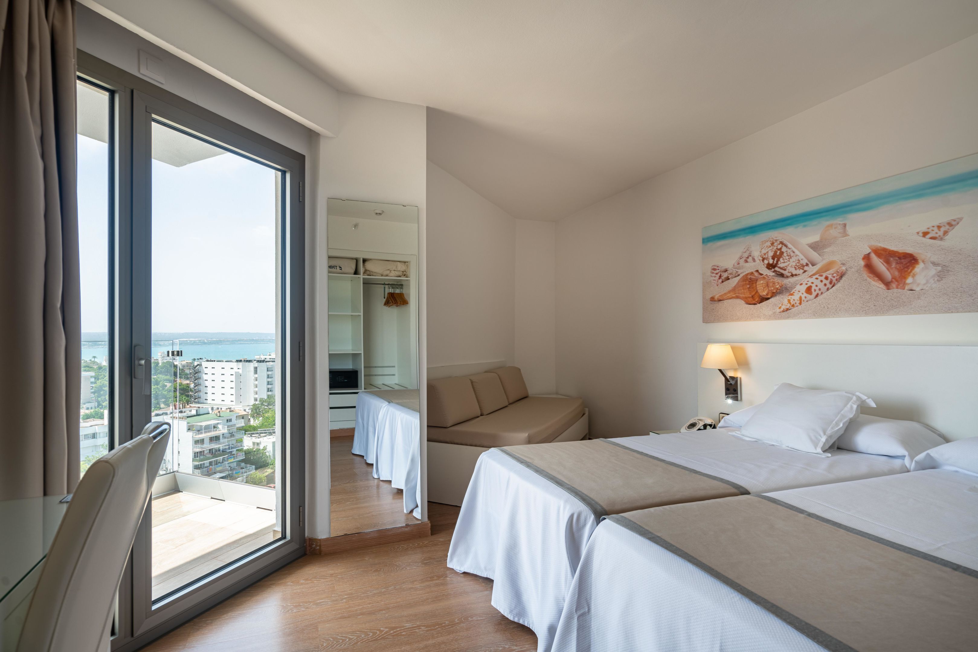 Een hotelkamer met een groot raam, een bank, twee bedden en een schilderij van schelpen.