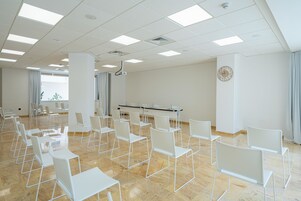 Un salón de conferencias con sillas y mesas blancas, un proyector y un reloj en la pared.