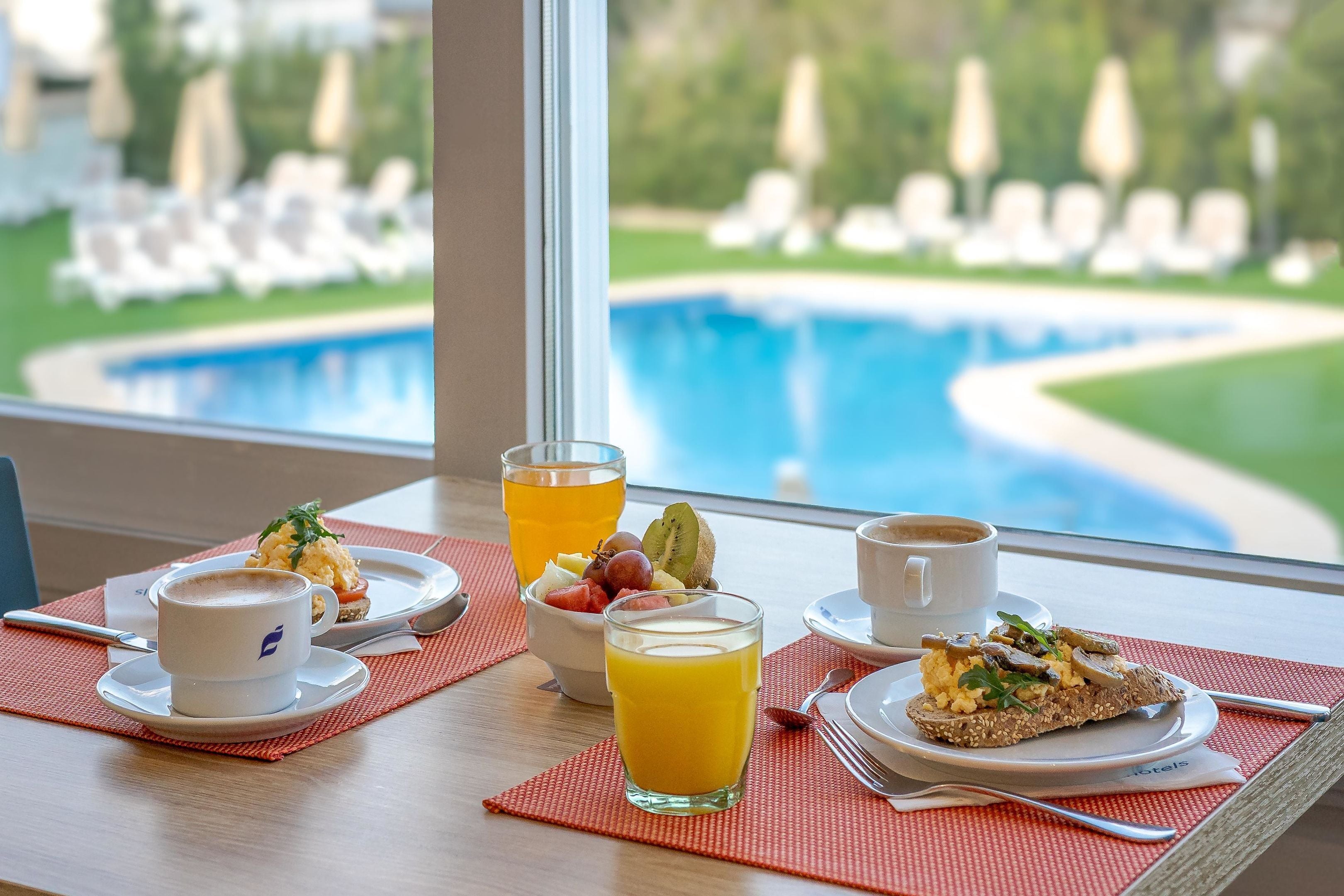 A breakfast setting with a pool view, including a cup of coffee, a glass of orange juice, and a plate with eggs and fruit.
