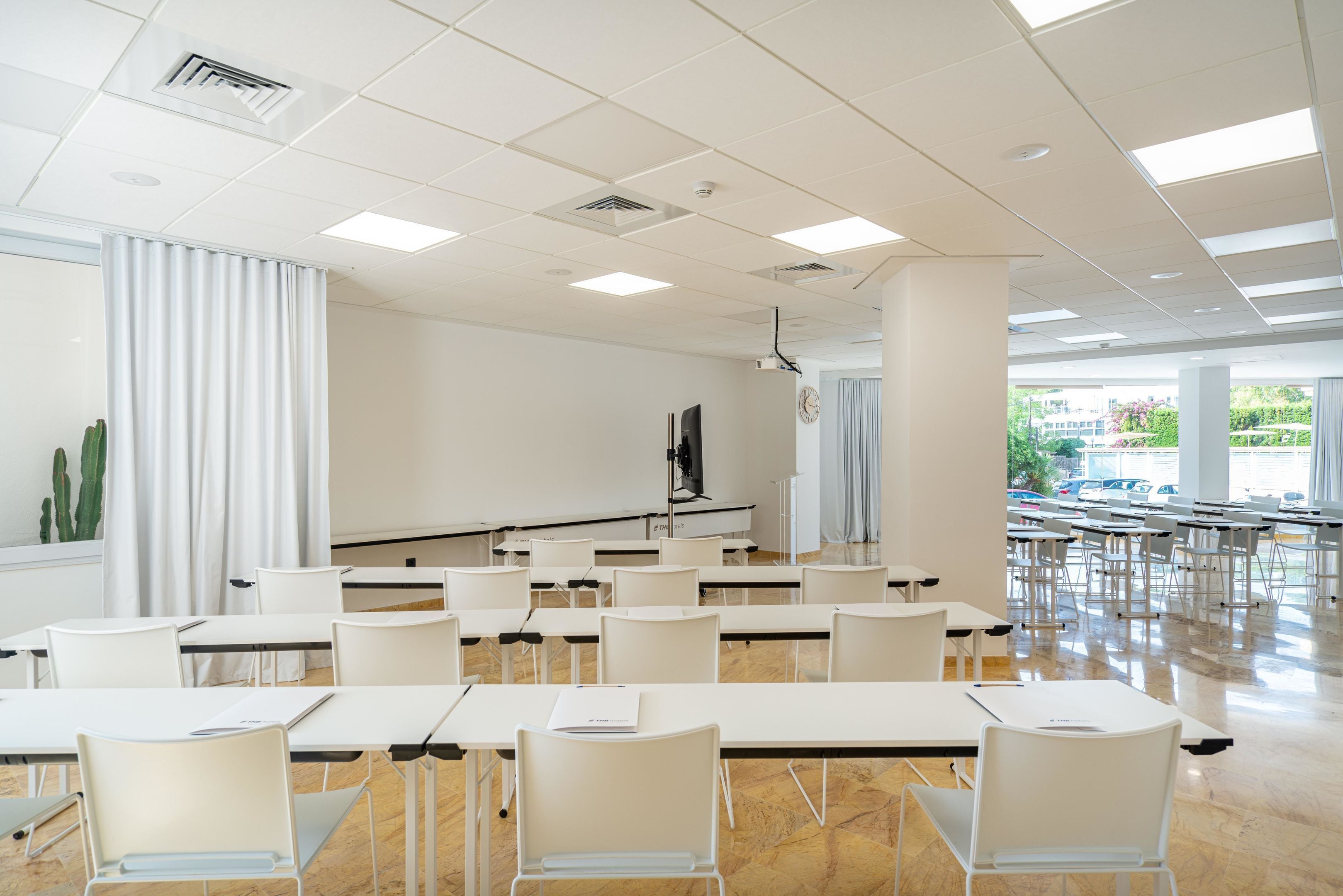 Un salón de conferencias con mesas y sillas blancas, un proyector y un ventanal con vistas a coches estacionados y vegetación.