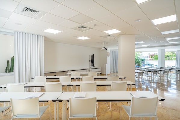 A conference room with white tables and chairs, a projector, and a large window with a view of parked cars and greenery outside.