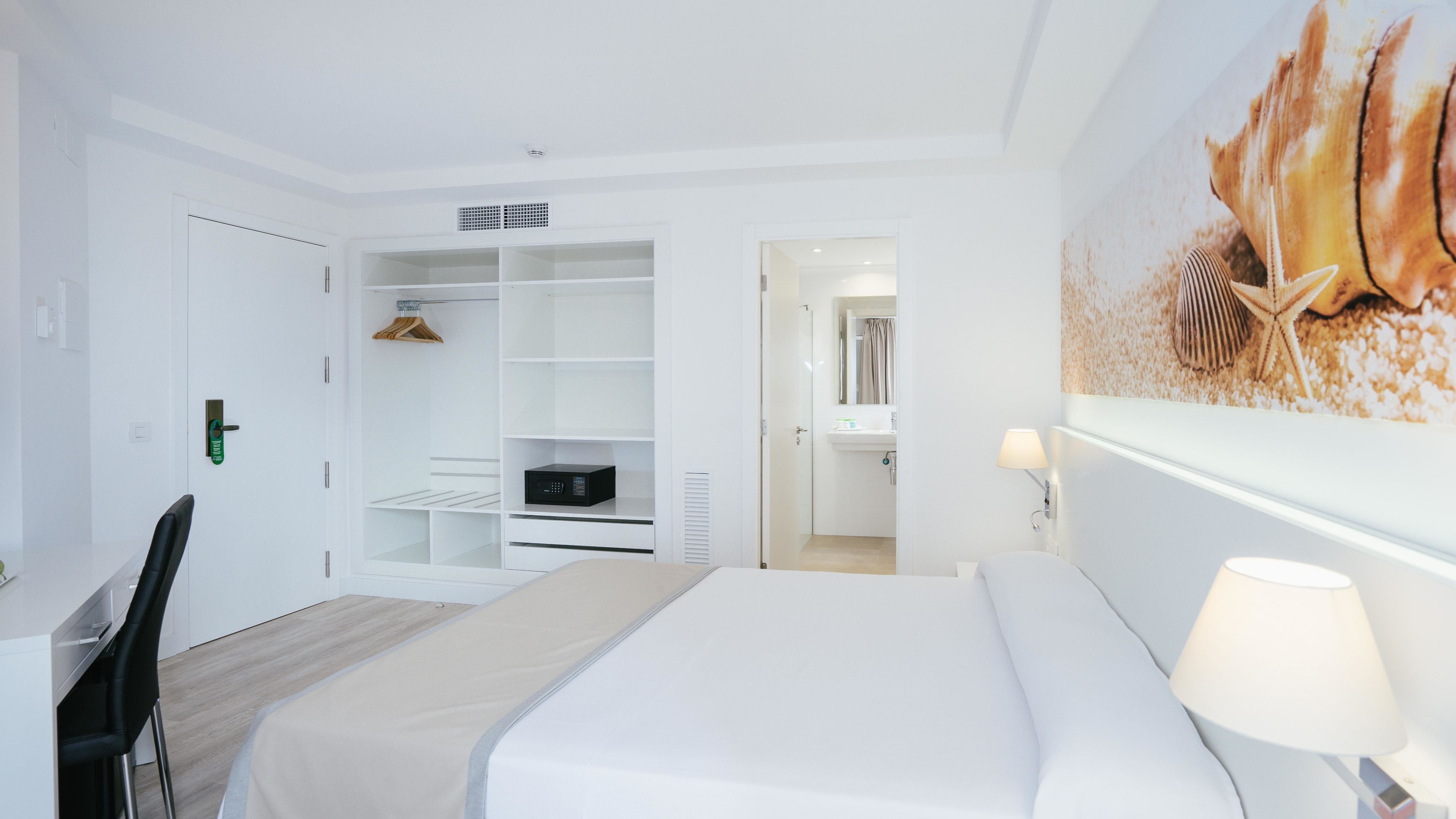 Habitación de hotel con cama, escritorio, silla, armario y baño visible a través de una puerta abierta.