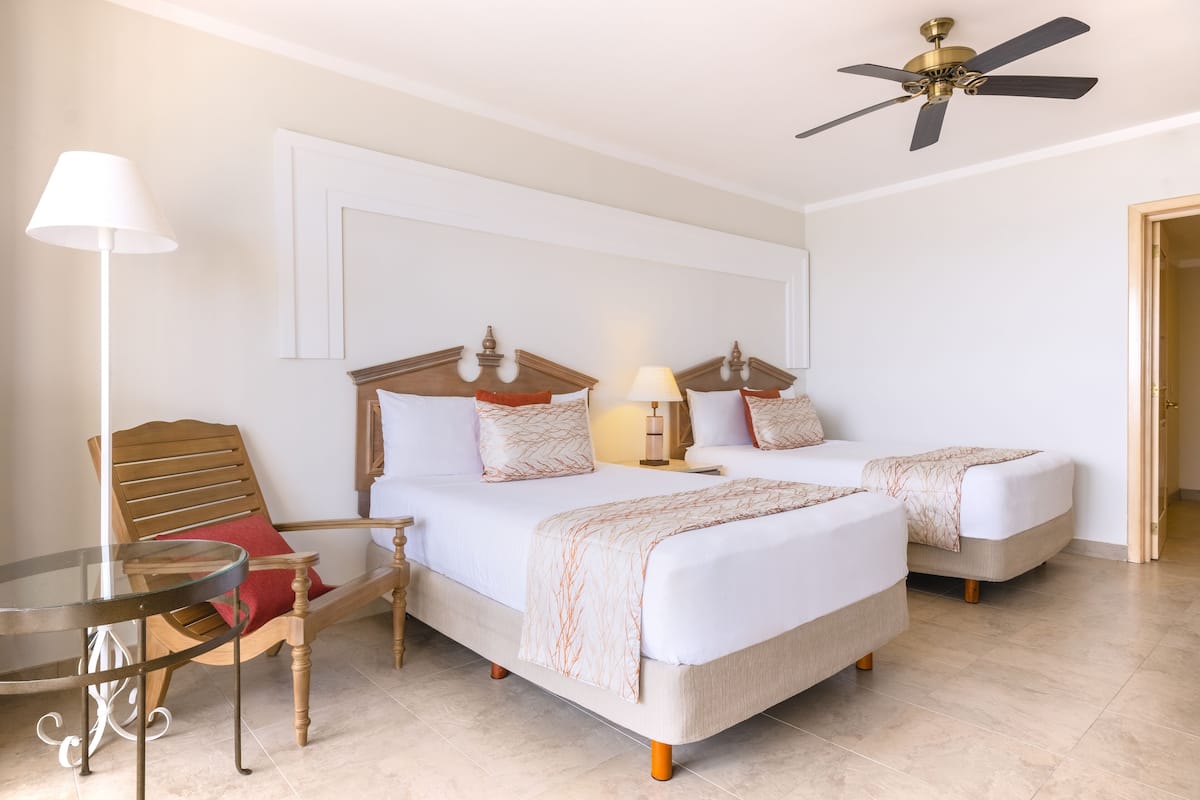 A hotel room with two beds, a wooden chair, a glass table, and a ceiling fan.