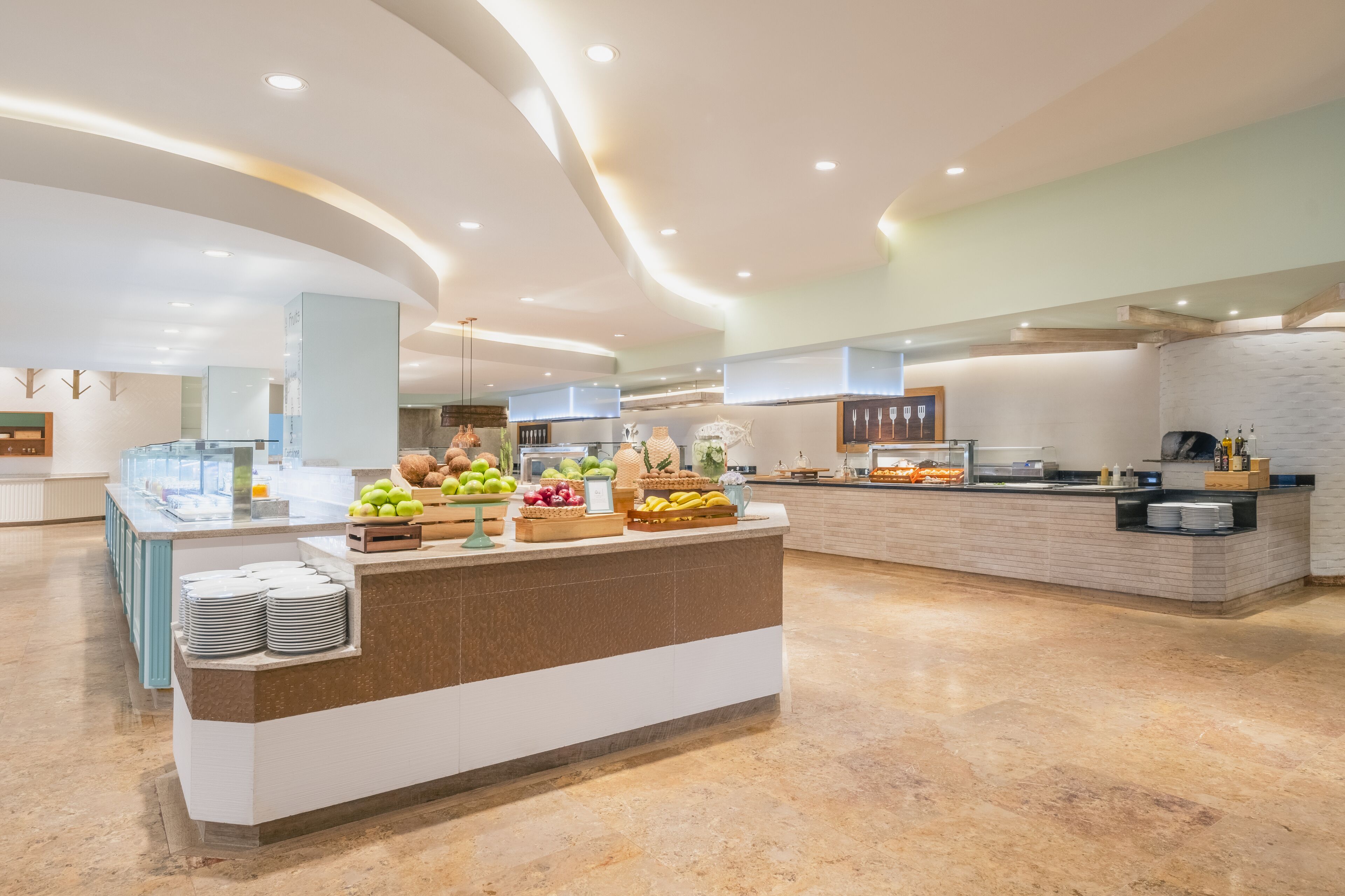 A modern hotel lobby with a central reception desk, a glass display case with fruits, and a buffet area with various food items.