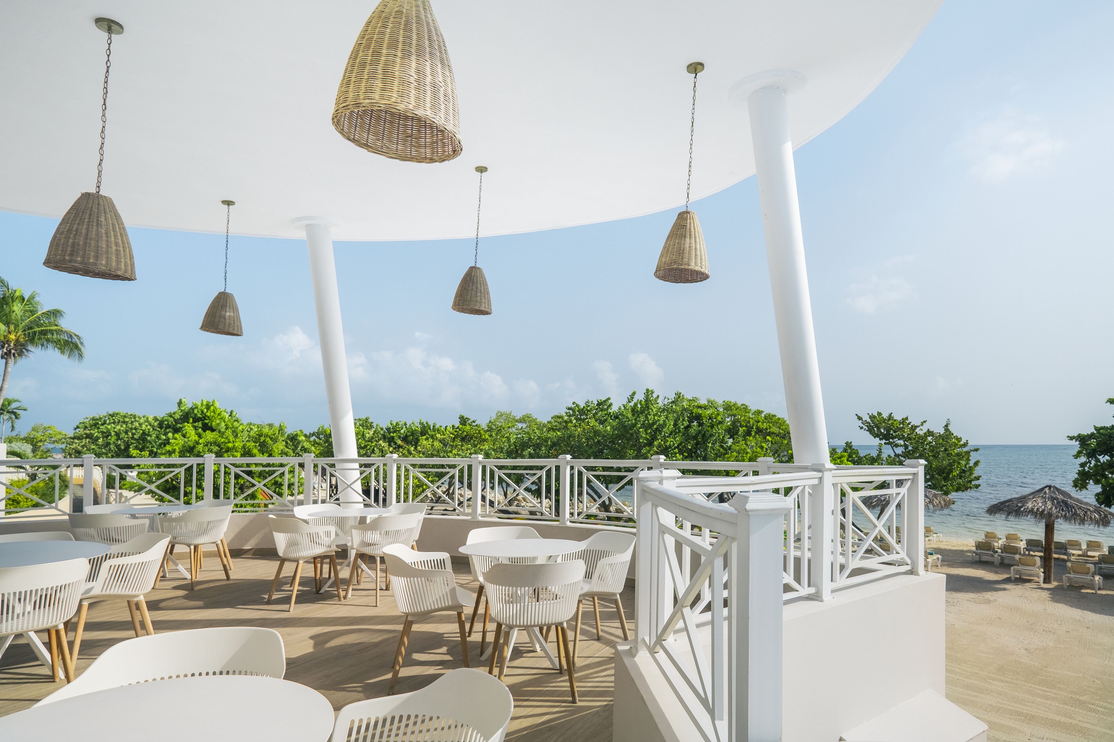An outdoor seating area with white chairs and tables, hanging wicker lamps, and a view of the ocean.