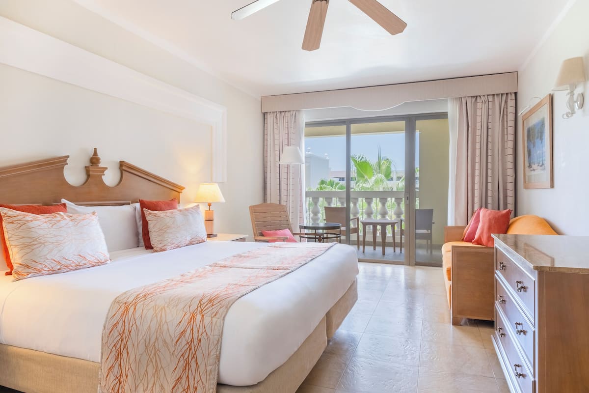 A hotel room with a large bed, a desk, a chair, and a balcony with a view of the outdoors.
