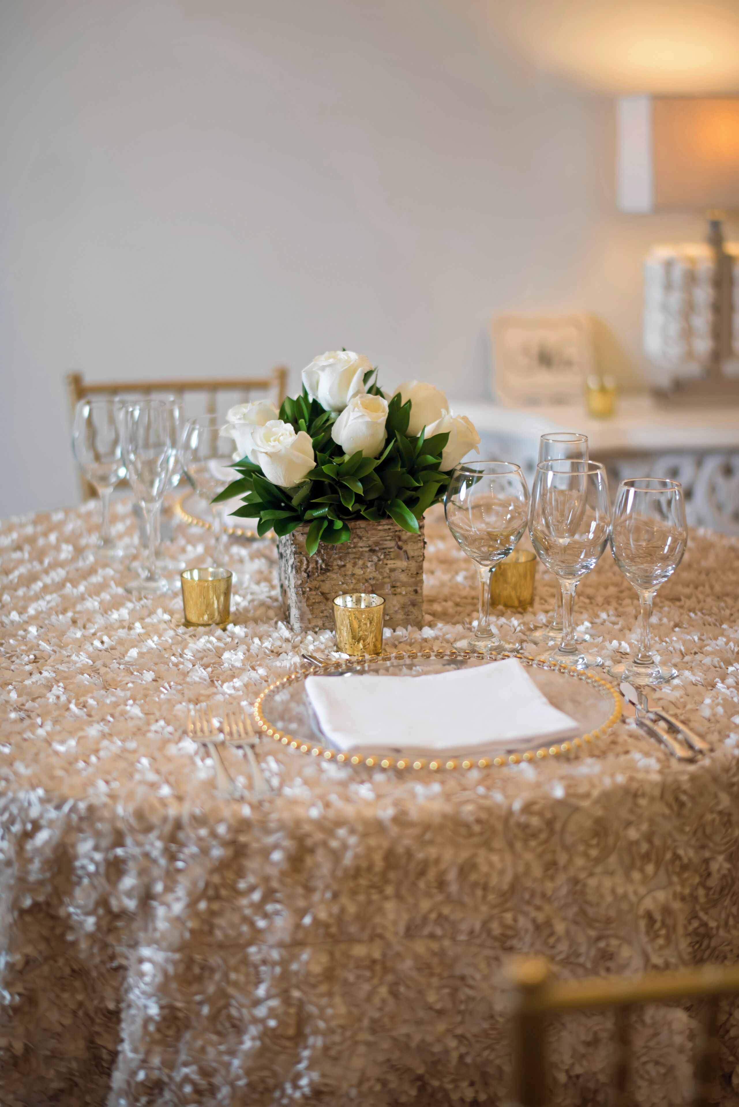 A table set with white flowers in a rustic vase, gold candle holders, and clear wine glasses.