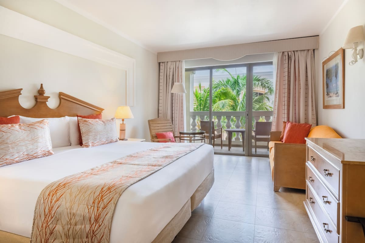 A hotel room with a large bed, a dresser, a desk with a chair, and a balcony with a view of palm trees.