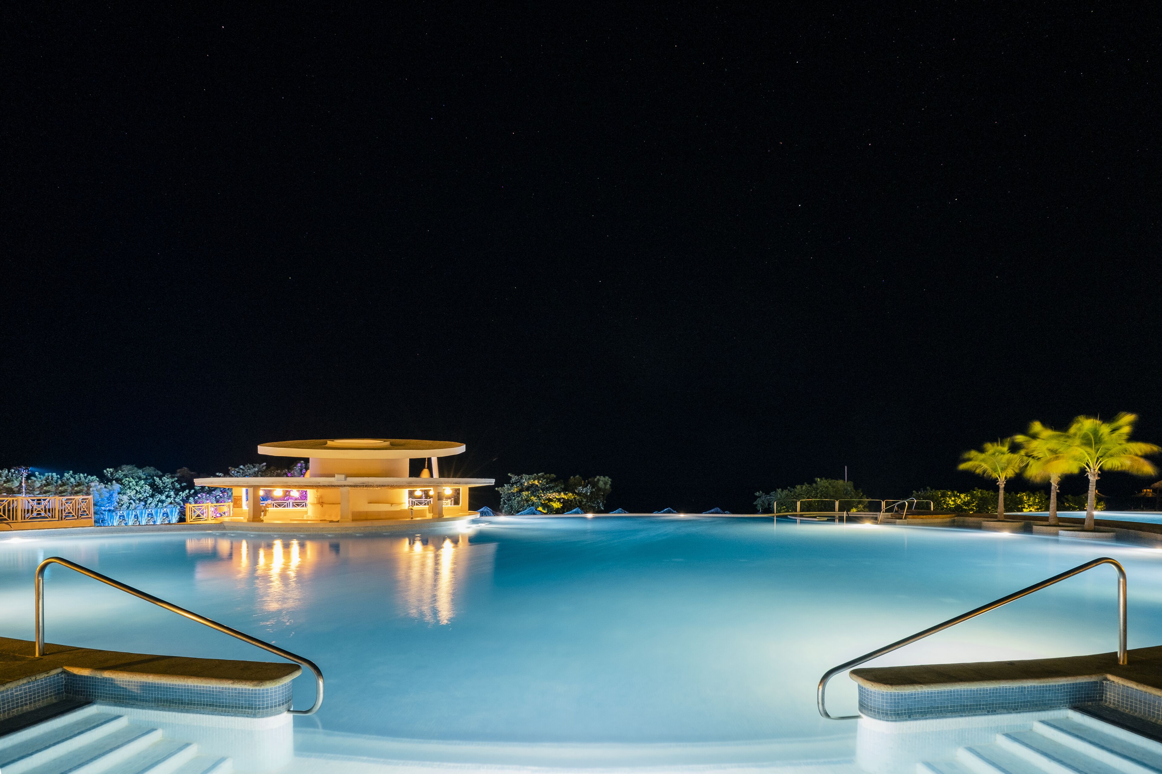 Une piscine nocturne avec un bâtiment circulaire et des palmiers.