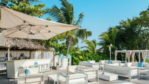 A deck with white furniture, a bar area, and palm trees in the background. - Iberostar Waves Rose Hall Beach - All Inclusive (Montego Bay)