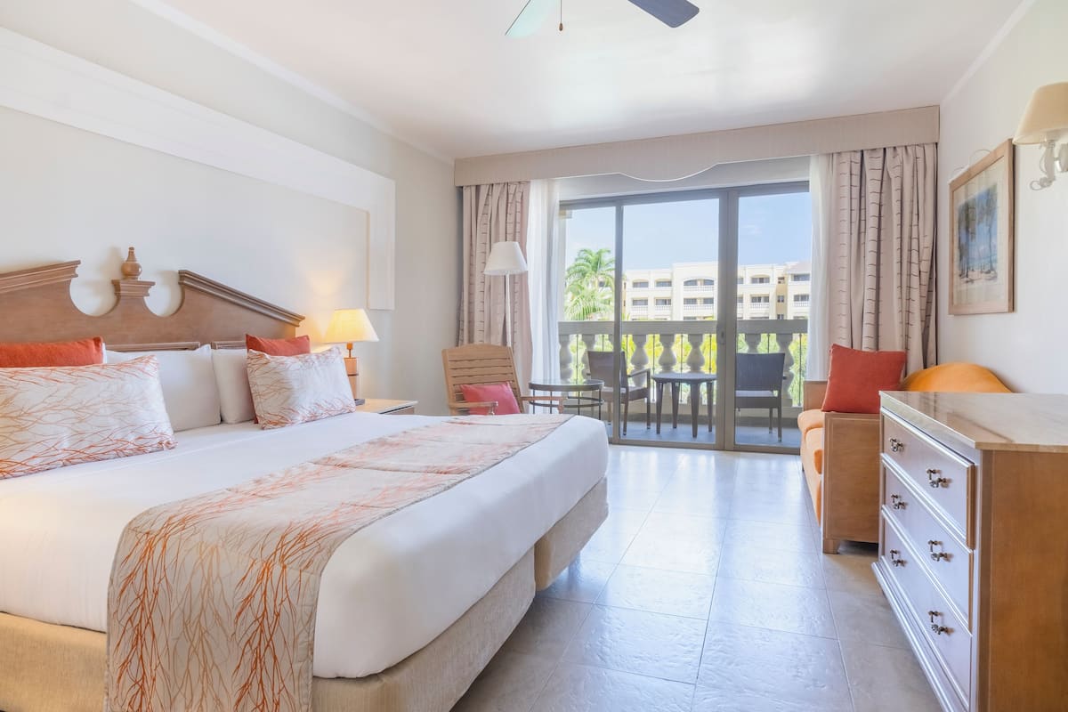 A hotel room with a large bed, a desk, a chair, and a balcony with a view of palm trees and buildings.