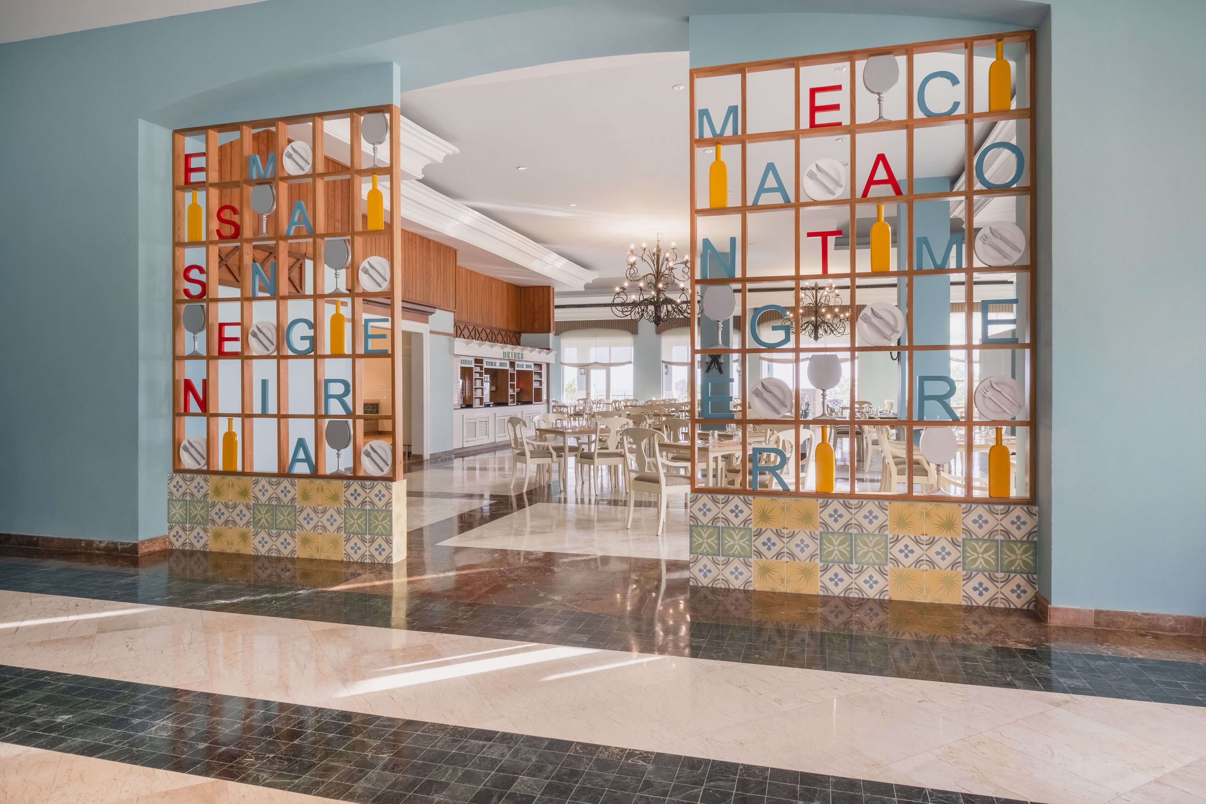 A hotel lobby with a tiled floor, a reception desk, and a decorative wall with letters and bottle silhouettes.