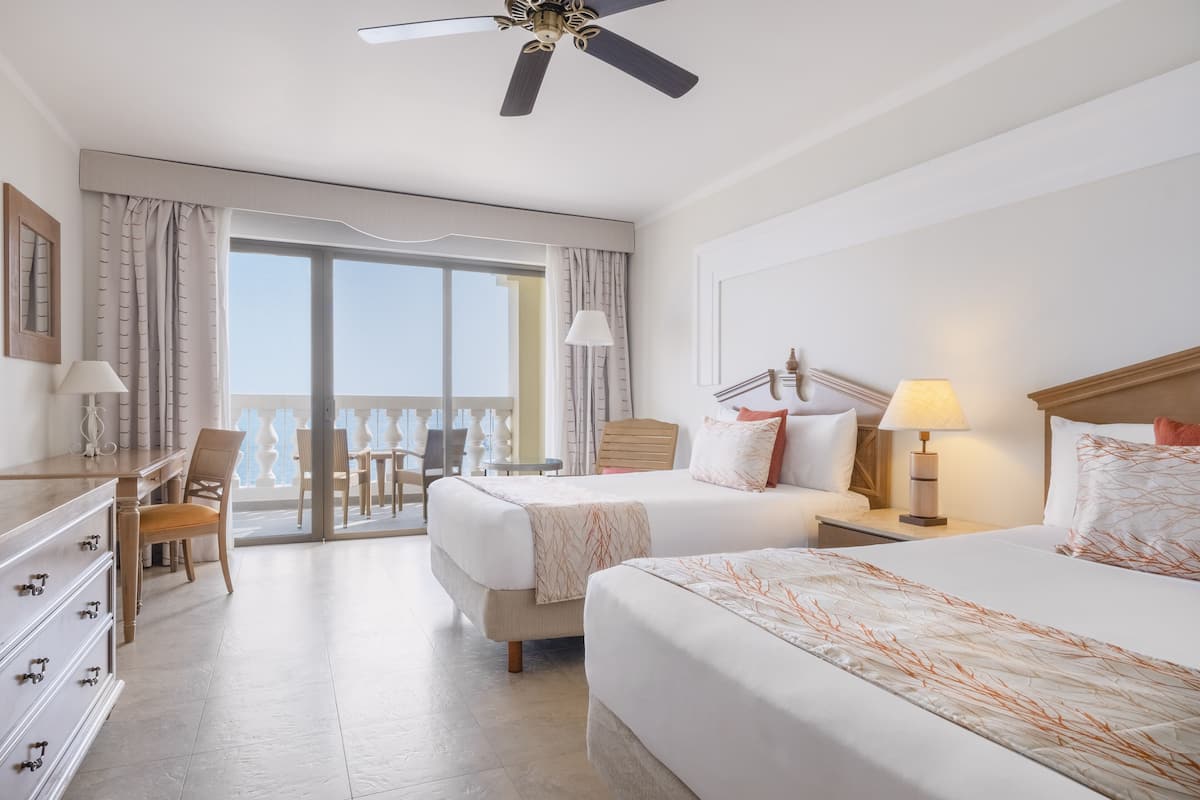 A hotel room with two beds, a desk, and a chair. There is a balcony with curtains and a view of the sky.