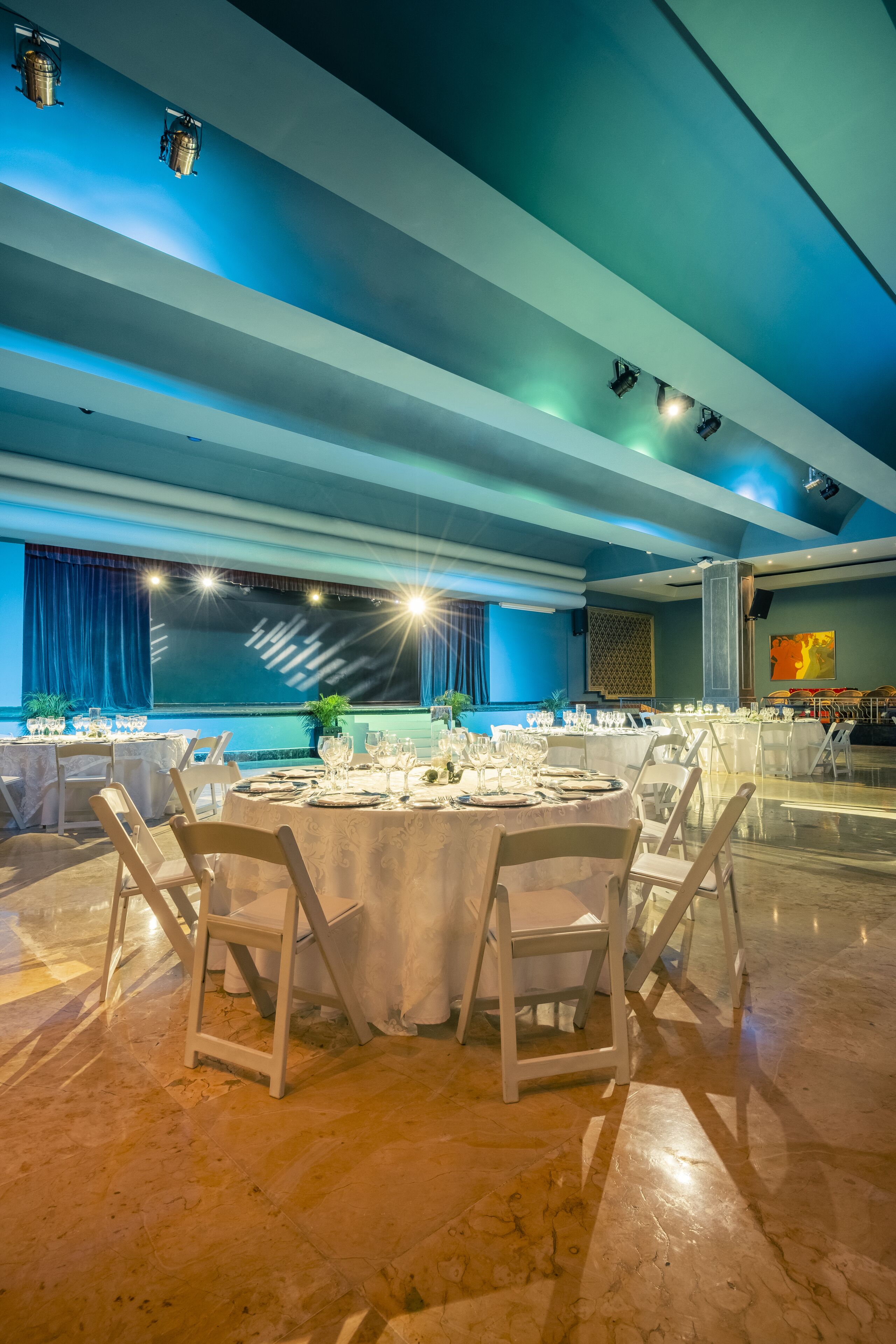 A spacious banquet hall with round tables set for dining, a high ceiling with blue lighting, and a large window with curtains.
