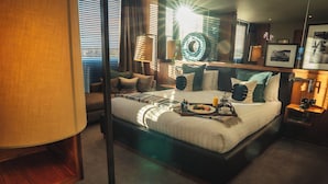 A hotel room with a large bed, a bedside table with a lamp, a tray with breakfast items, and a window with blinds. - Aviator Hampshire (Farnborough)