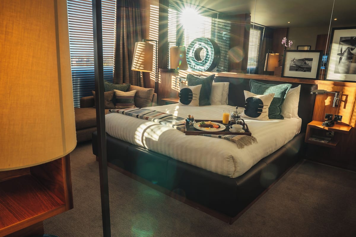 A hotel room with a large bed, a bedside table with a lamp, a tray with breakfast items, and a window with blinds.