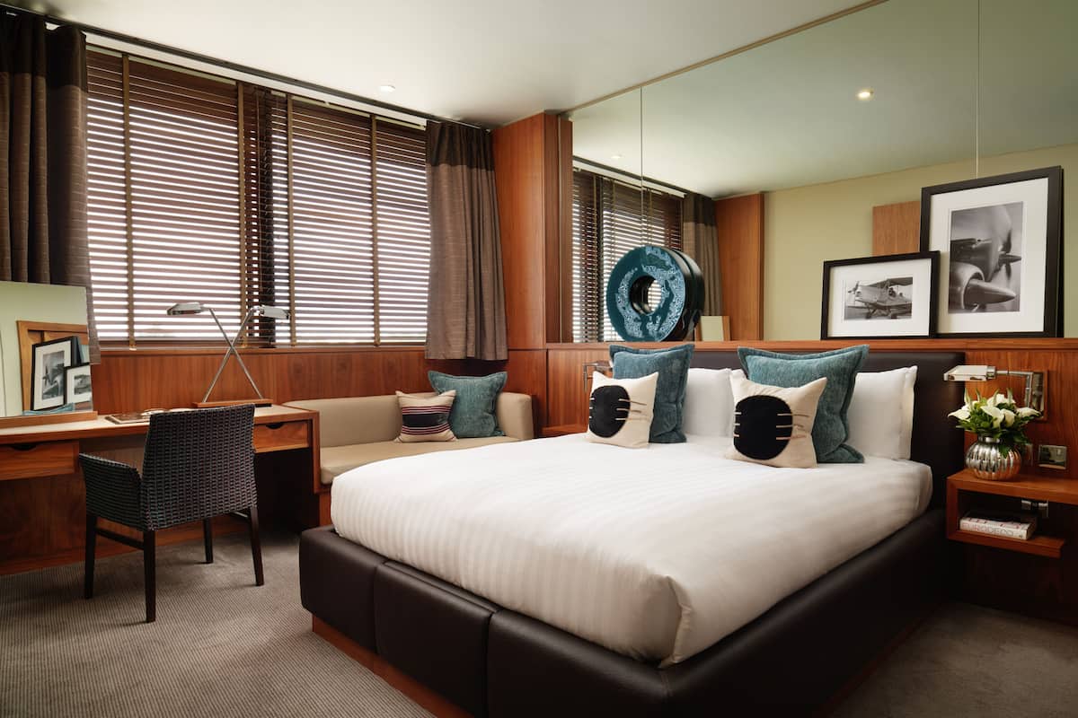 A modern hotel room with a large bed, a desk, and a chair. There are curtains on the windows, a wall-mounted lamp, and framed pictures on the wall.