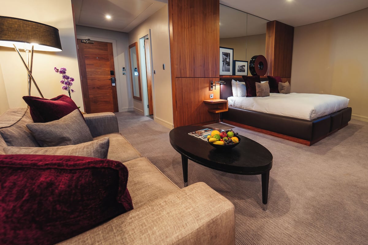 A modern hotel room with a sofa, a coffee table with fruit, a bed, and a wooden accent wall.