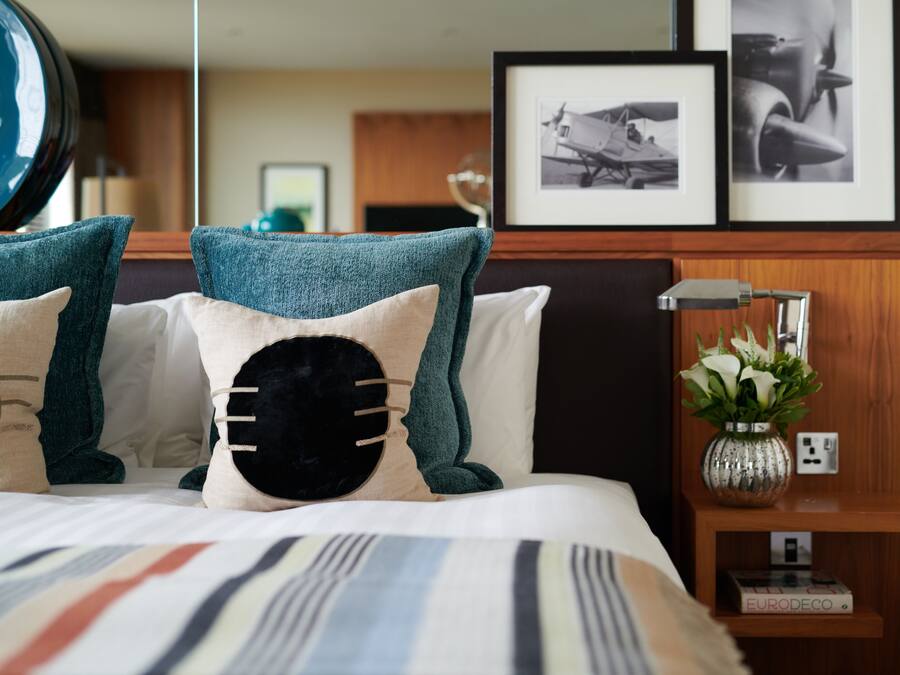 A neatly made bed with striped bedding, a bedside table with a lamp, a framed picture of an airplane, and a small shelf with a plant and a book.