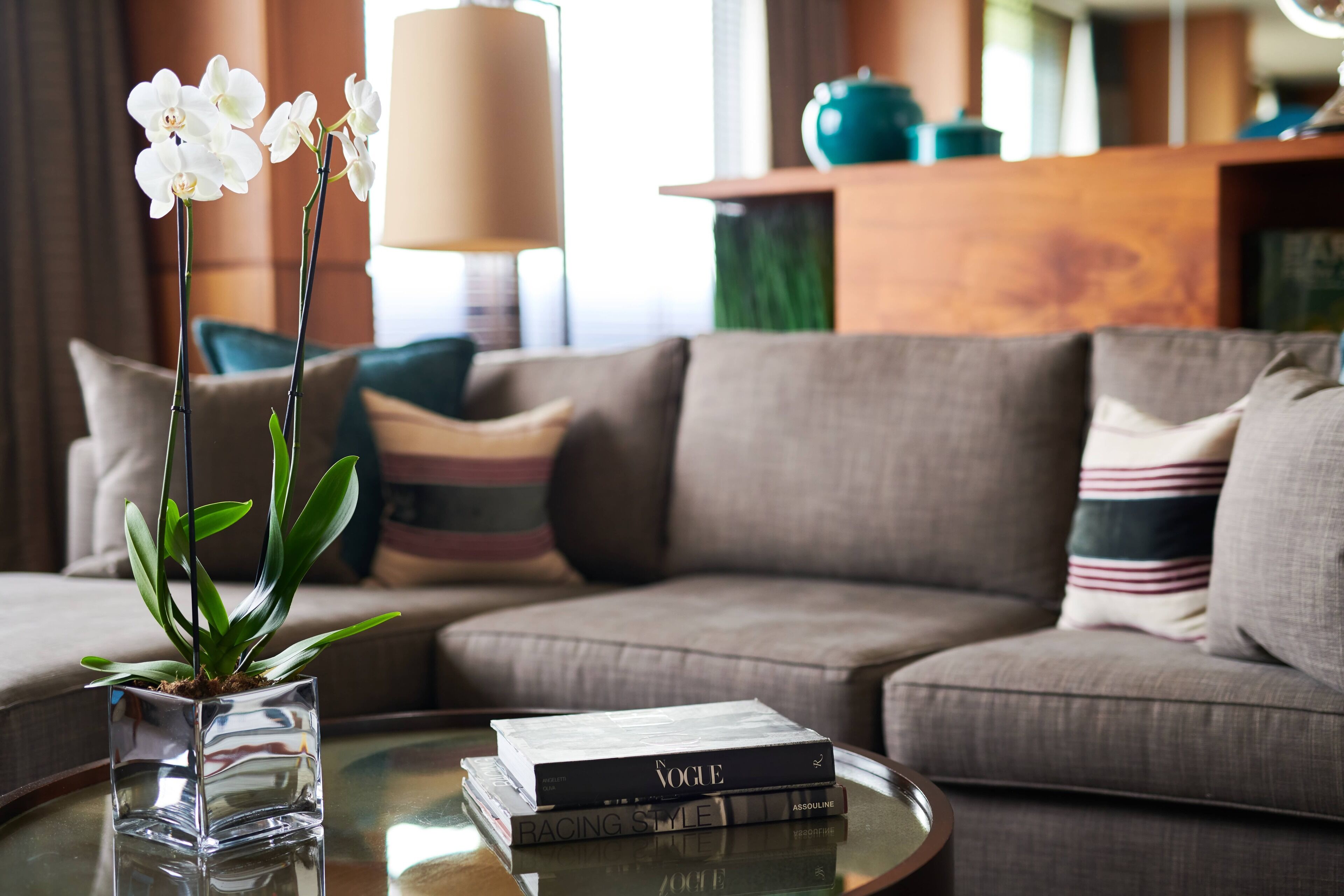 En moderne stue med en sofa, et glasbord, en vase med hvide orkidéer og en stak bøger med titlen 'Vogue'.