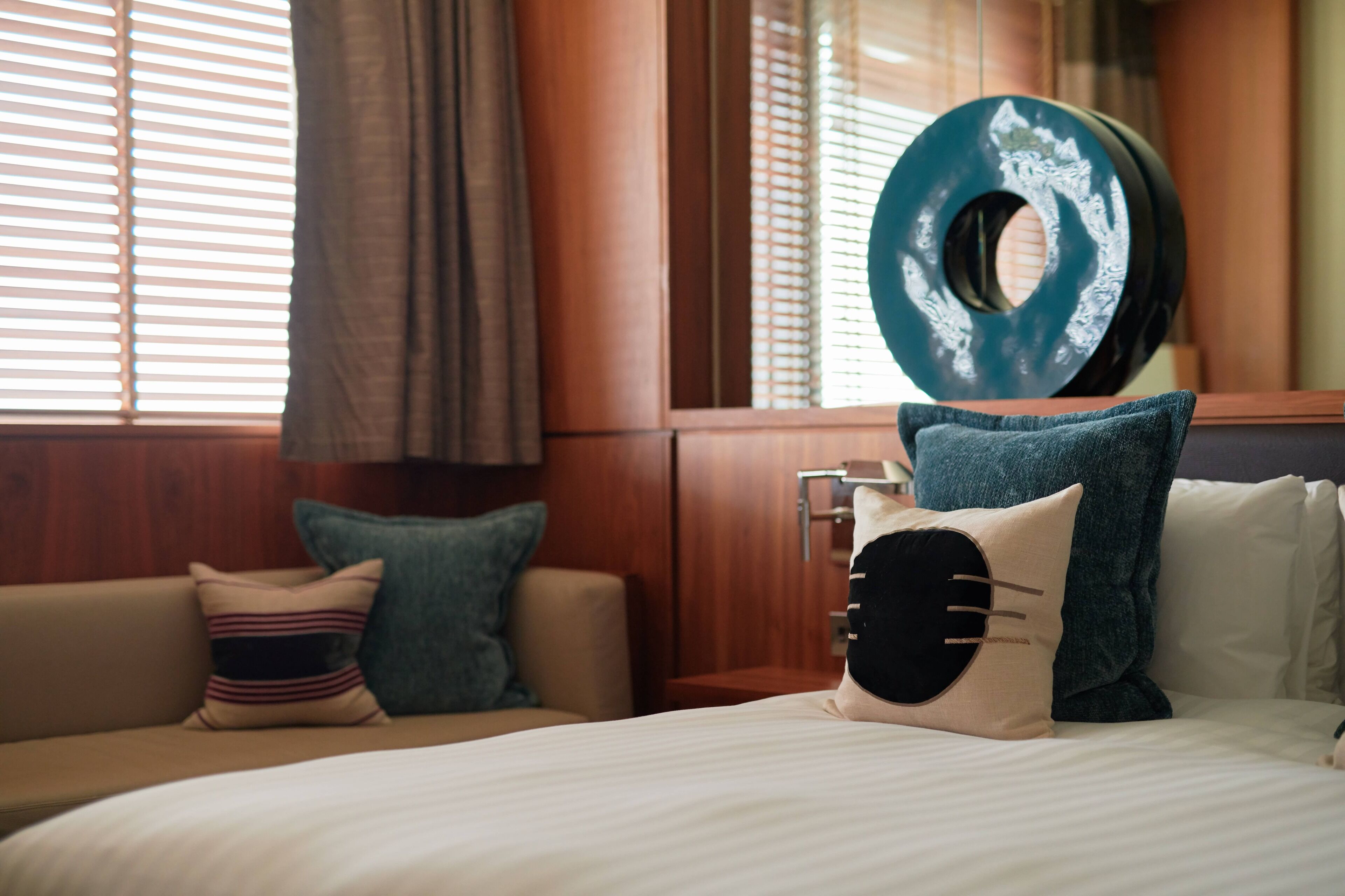 Une chambre d’hôtel avec un lit, une tête de lit en bois, une fenêtre avec des stores et un grand miroir décoratif.