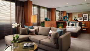 A modern hotel room with a sectional sofa, a round coffee table, a bed with a headboard, a mirror, and a bookshelf. - Aviator Hampshire (Farnborough)