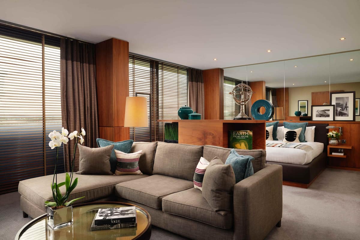 A modern hotel room with a sectional sofa, a round coffee table, a bed with a headboard, a mirror, and a bookshelf.