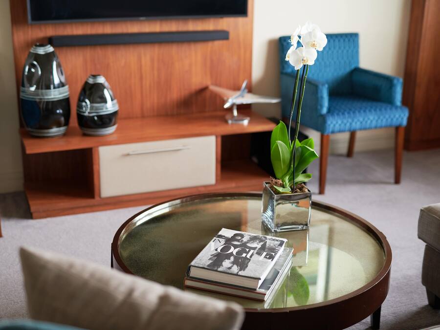A living room with a glass coffee table, a magazine, a vase with flowers, a blue armchair, and a wooden entertainment unit.