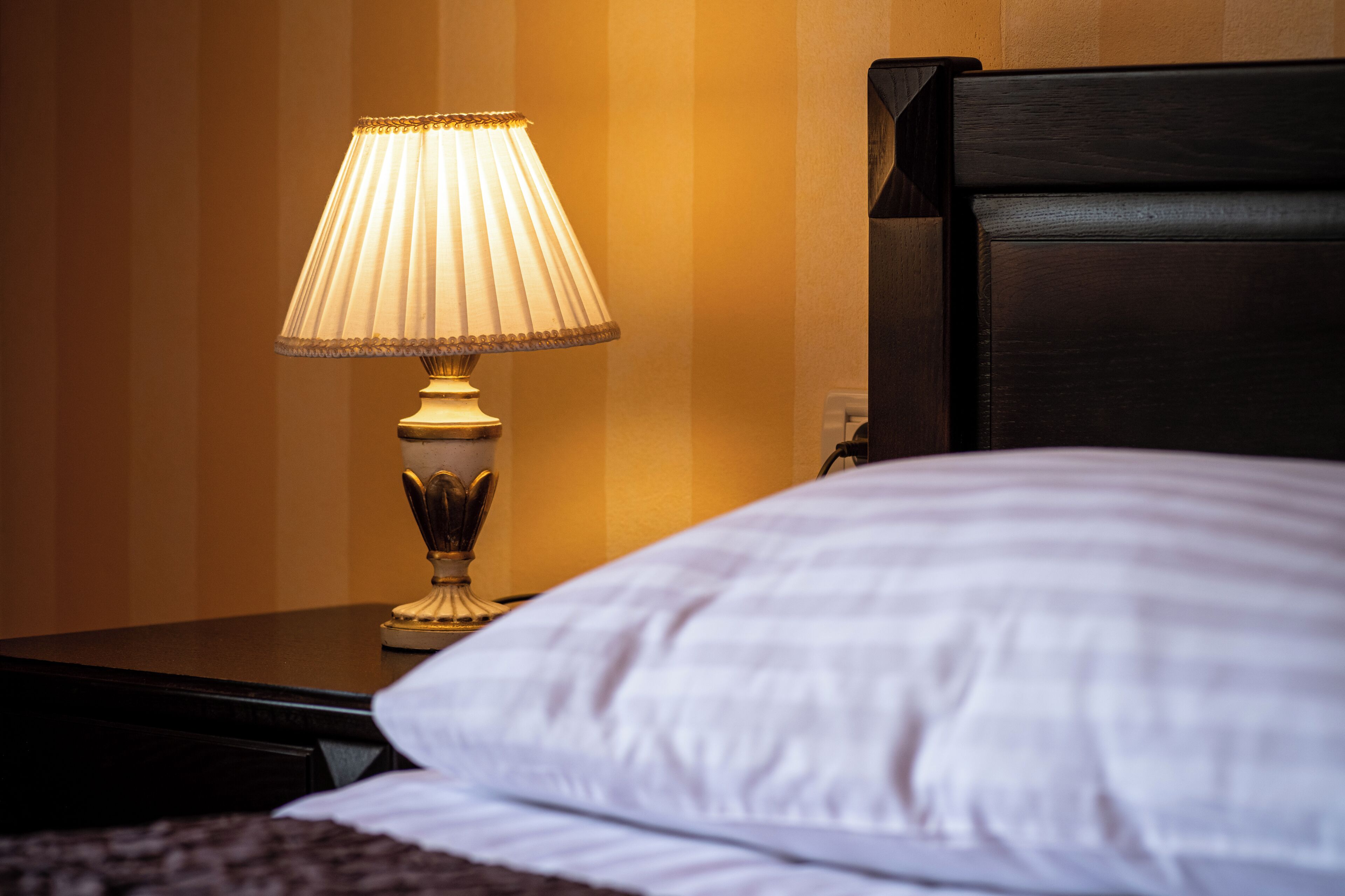 Cama com travesseiro branco e mesa de cabeceira com abajur.
