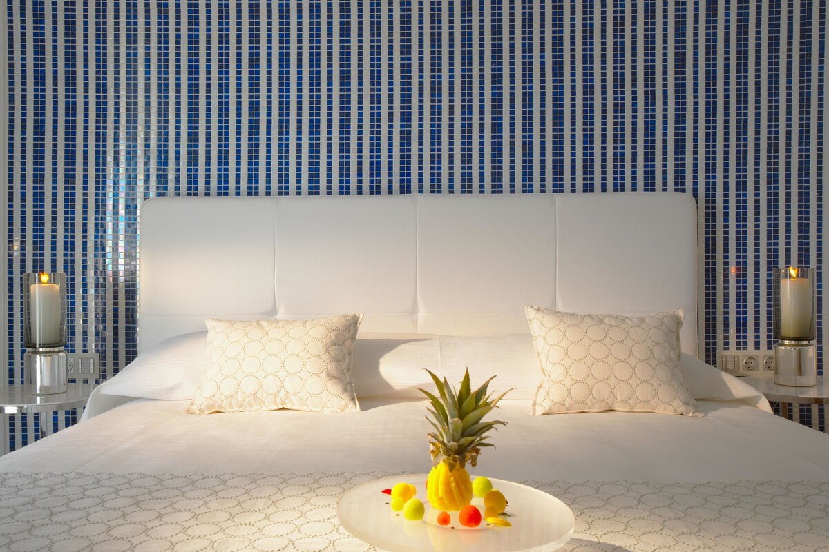 Une chambre d’hôtel avec un grand lit, deux oreillers blancs, un ananas et un plateau de fruits posés sur le lit.
