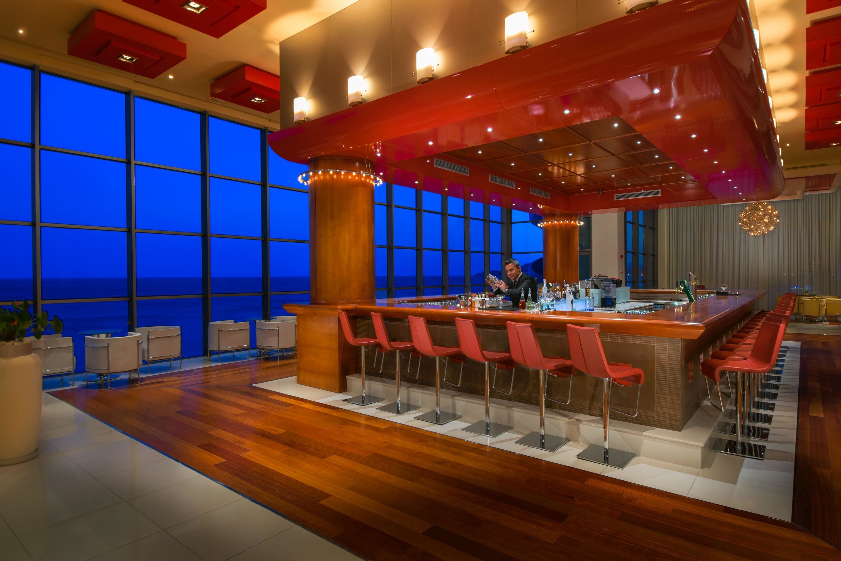 A modern bar area with a long counter, multiple red bar stools, and a view of the ocean through large windows.