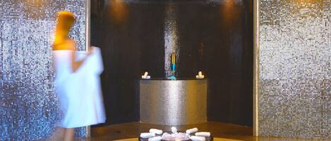 A spa area with a circular hot tub surrounded by candles, a tiled wall, and a person in a robe.