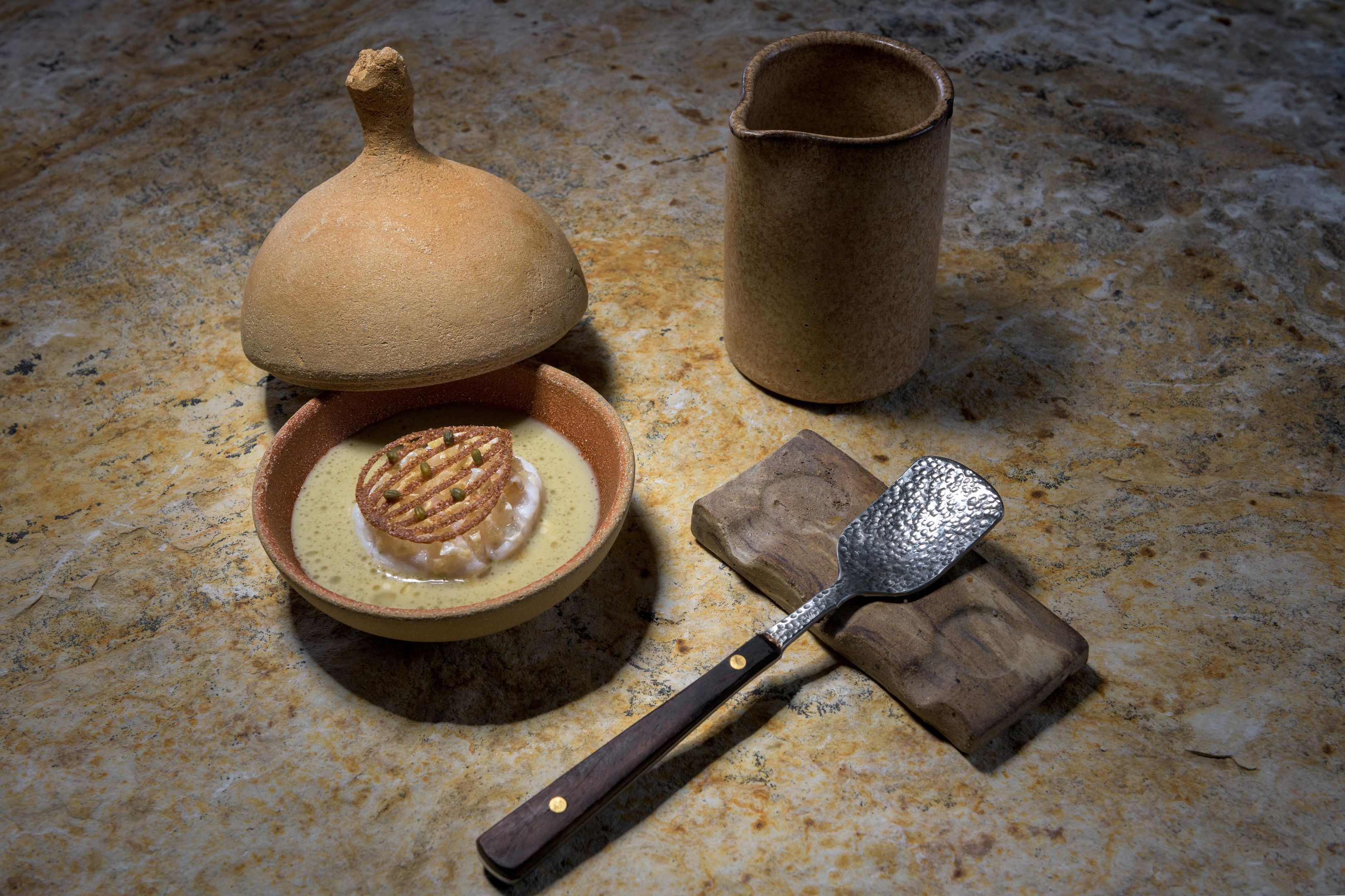 A clay pot with a lid, a bowl with a food item, a metal spatula, and a wooden cutting board on a textured surface.