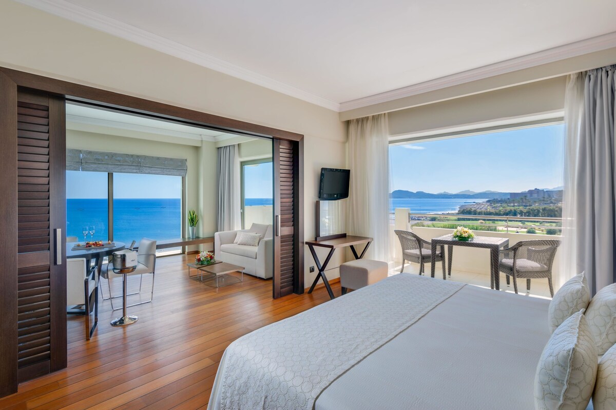 Une chambre d’hôtel avec un grand lit, un balcon donnant sur la mer, un coin repas avec une table et des chaises, et un téléviseur posé sur un meuble.