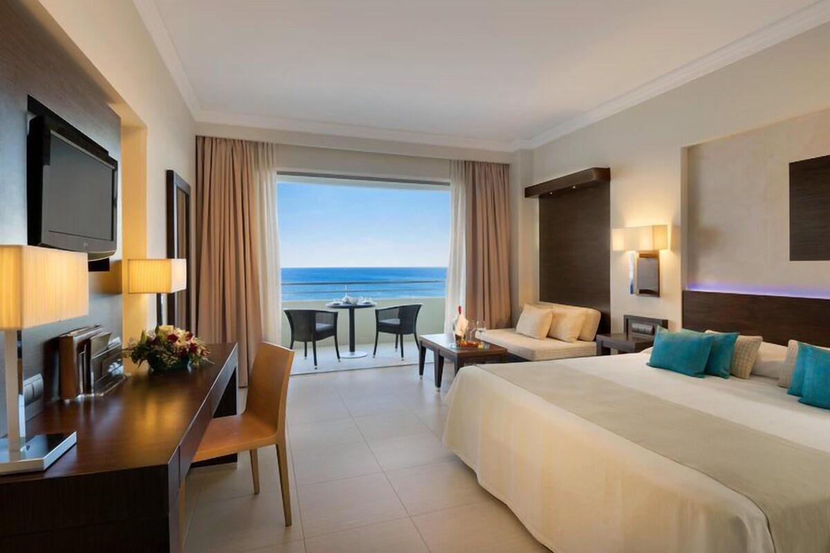 Una habitación de hotel con cama, escritorio, televisor, vista al océano y balcón.