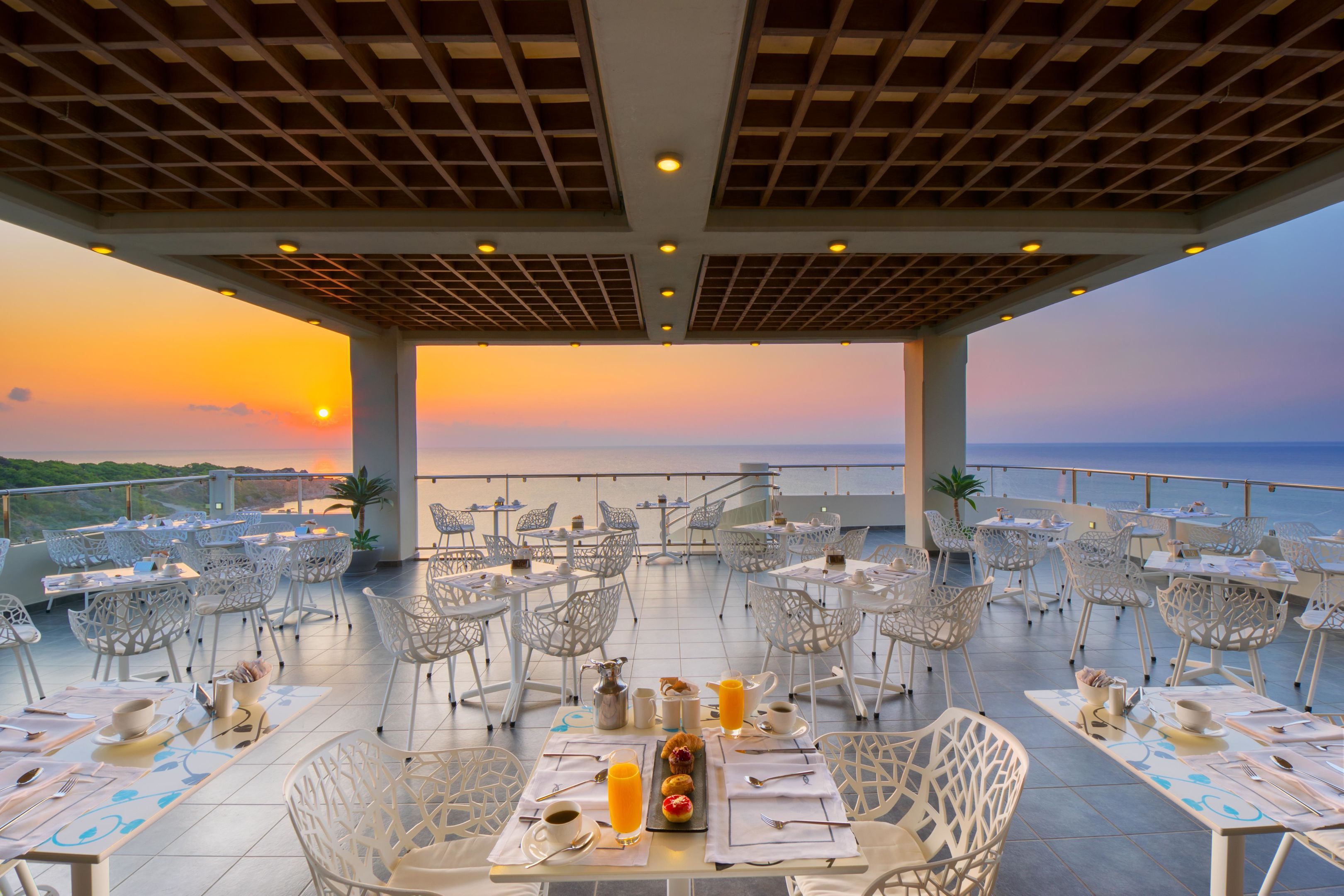 An outdoor dining area with tables and chairs, a sunset view, and a pool in the background.