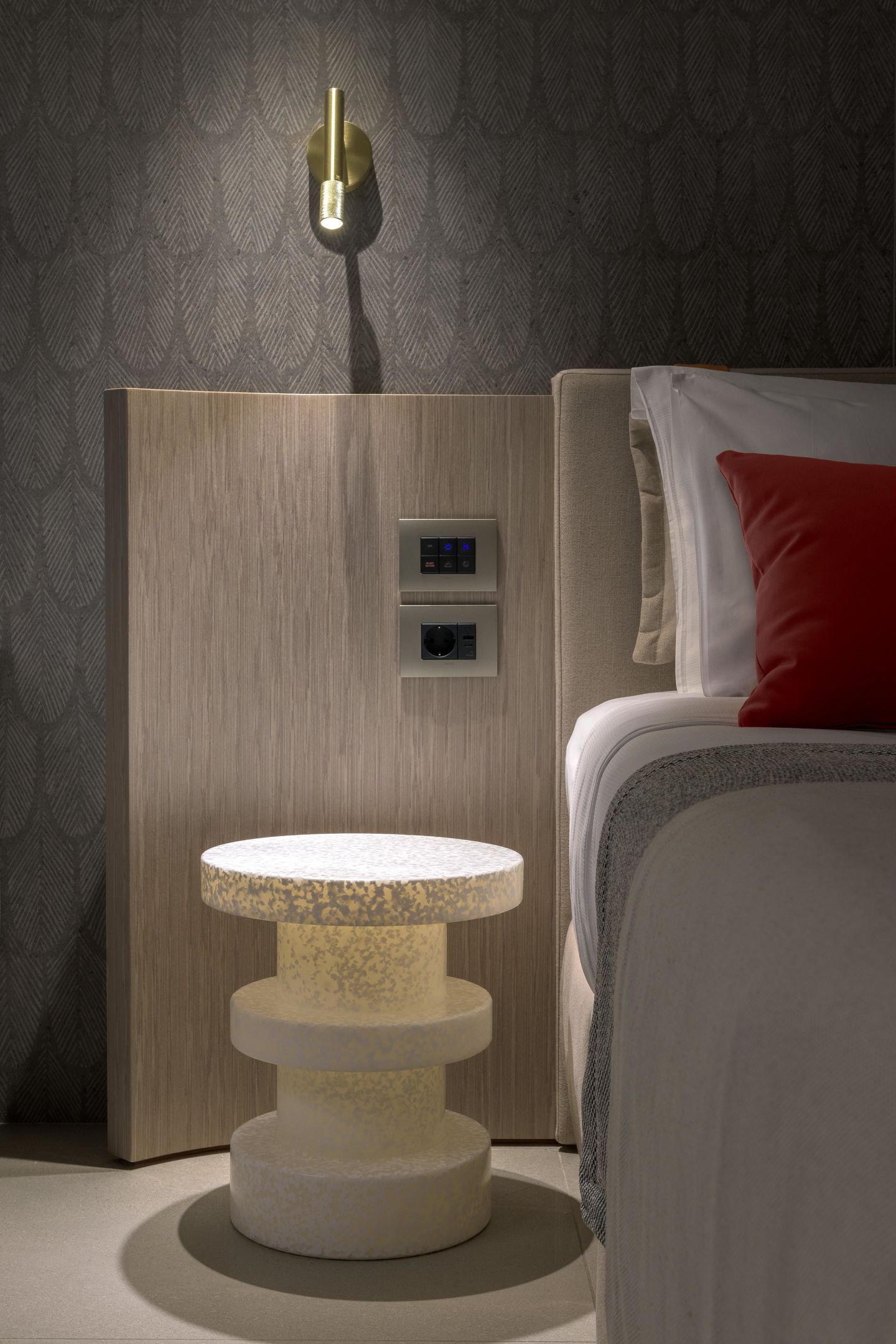 A modern hotel room with a wooden headboard, a bedside table with a lamp, a control panel, and a bed with a red pillow.
