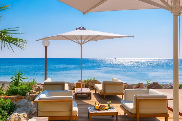 A wooden deck with white cushioned chairs and a table, overlooking a beach with clear blue water and a sailboat in the distance. - Elysium Resort & Spa (Rhodes)