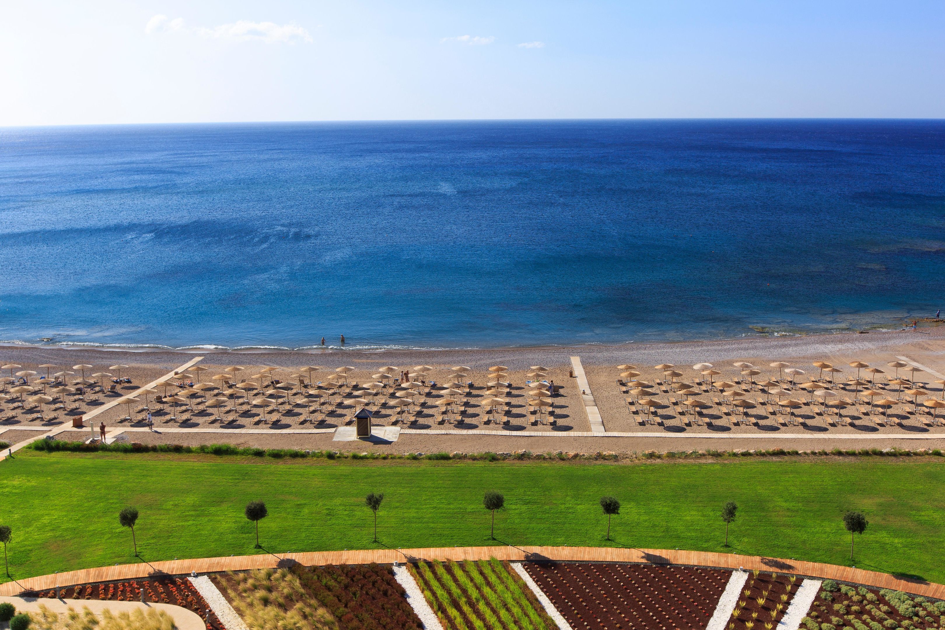 Vista aérea de un complejo turístico costero con zona de playa, hileras de sombrillas y jardines ajardinados.