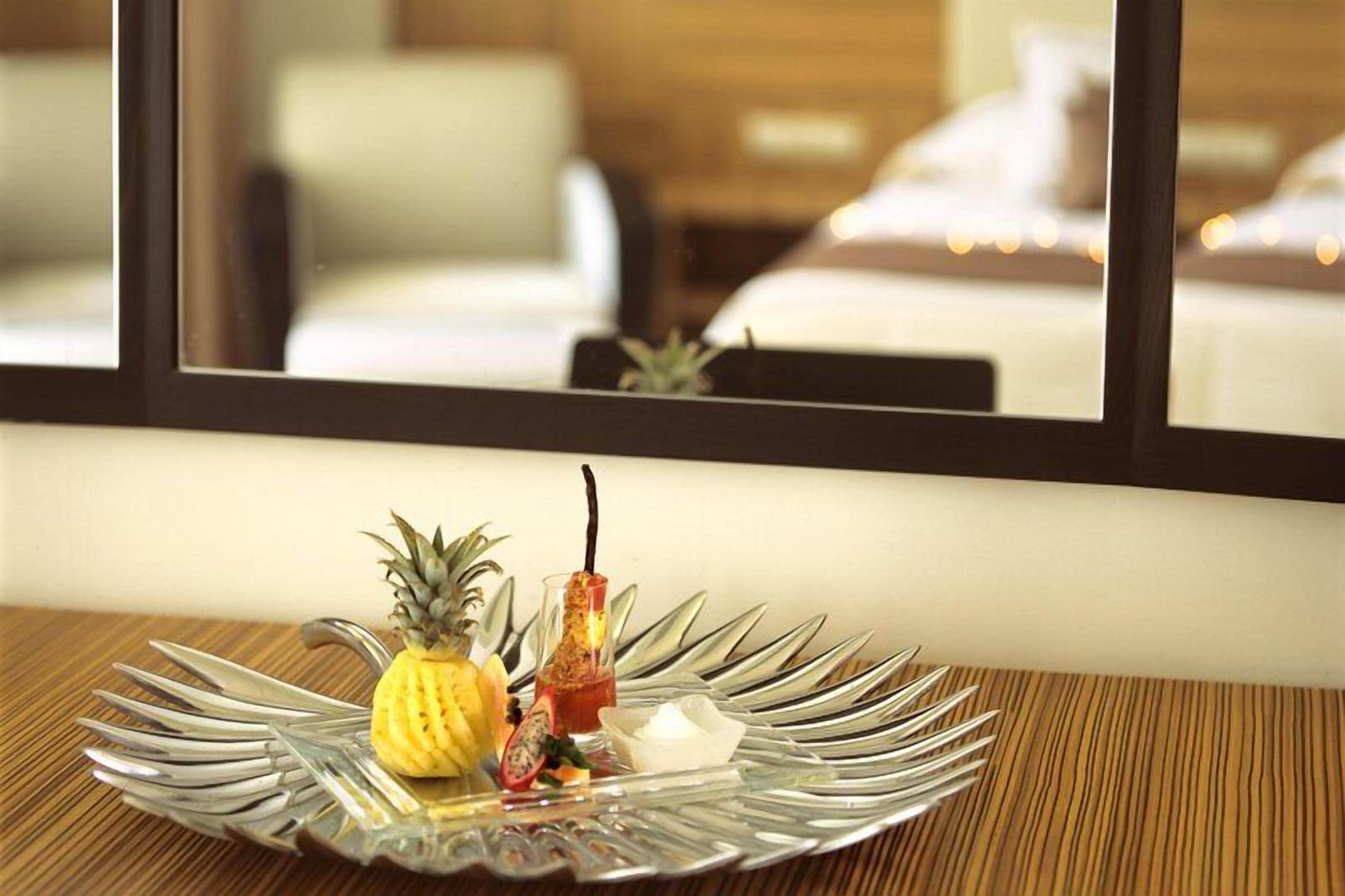 Una mesa de comedor con una bandeja de frutas y un vaso de bebida, una ventana con vista a una cama y una planta, y un suelo de madera.
