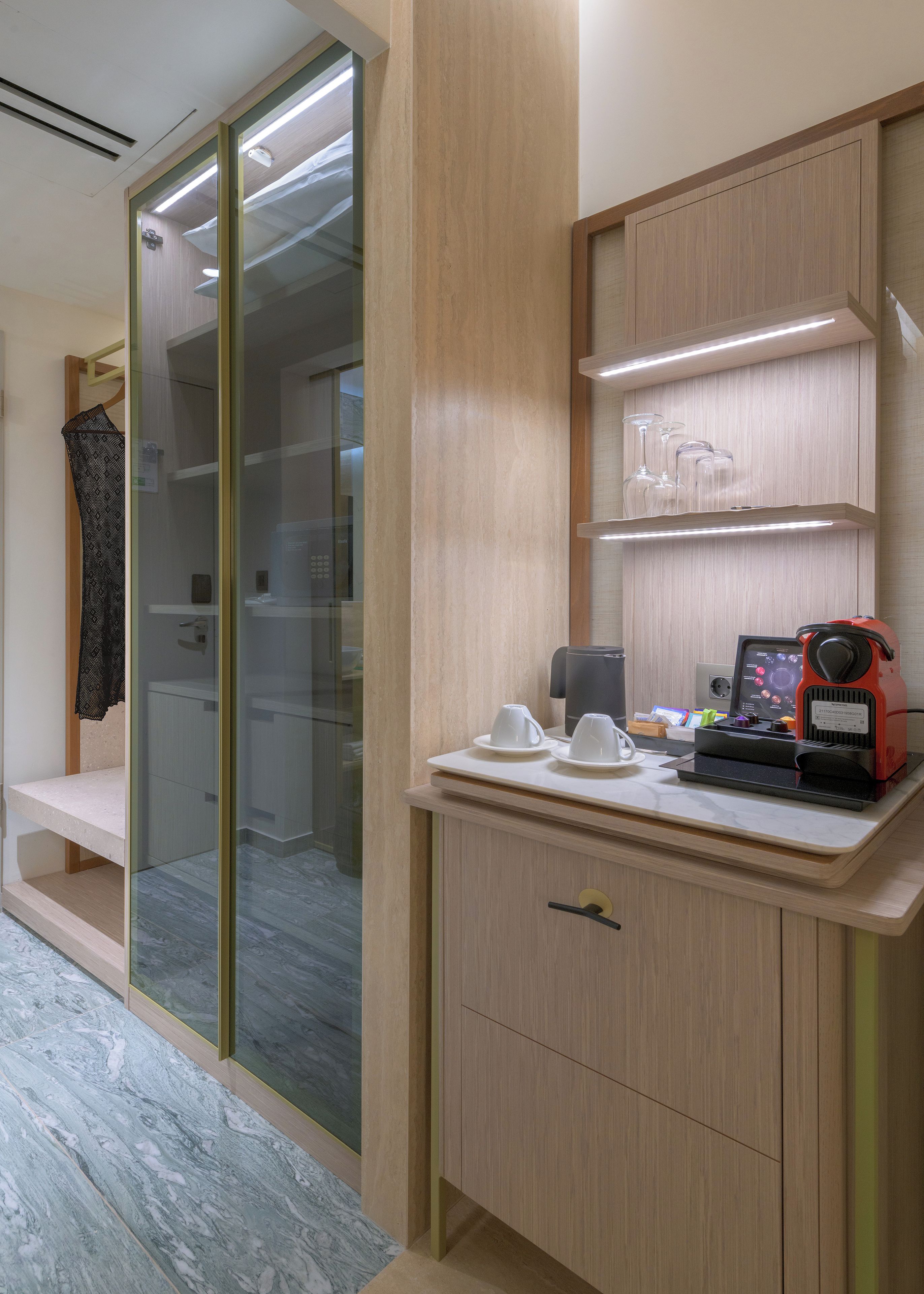 Una habitación de hotel moderna con un vestidor acristalado, un tocador de madera con una cafetera y un piso de baldosas de mármol.