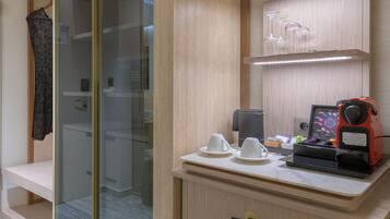 Una habitación de hotel moderna con un vestidor acristalado, un tocador de madera con una cafetera y un piso de baldosas de mármol.