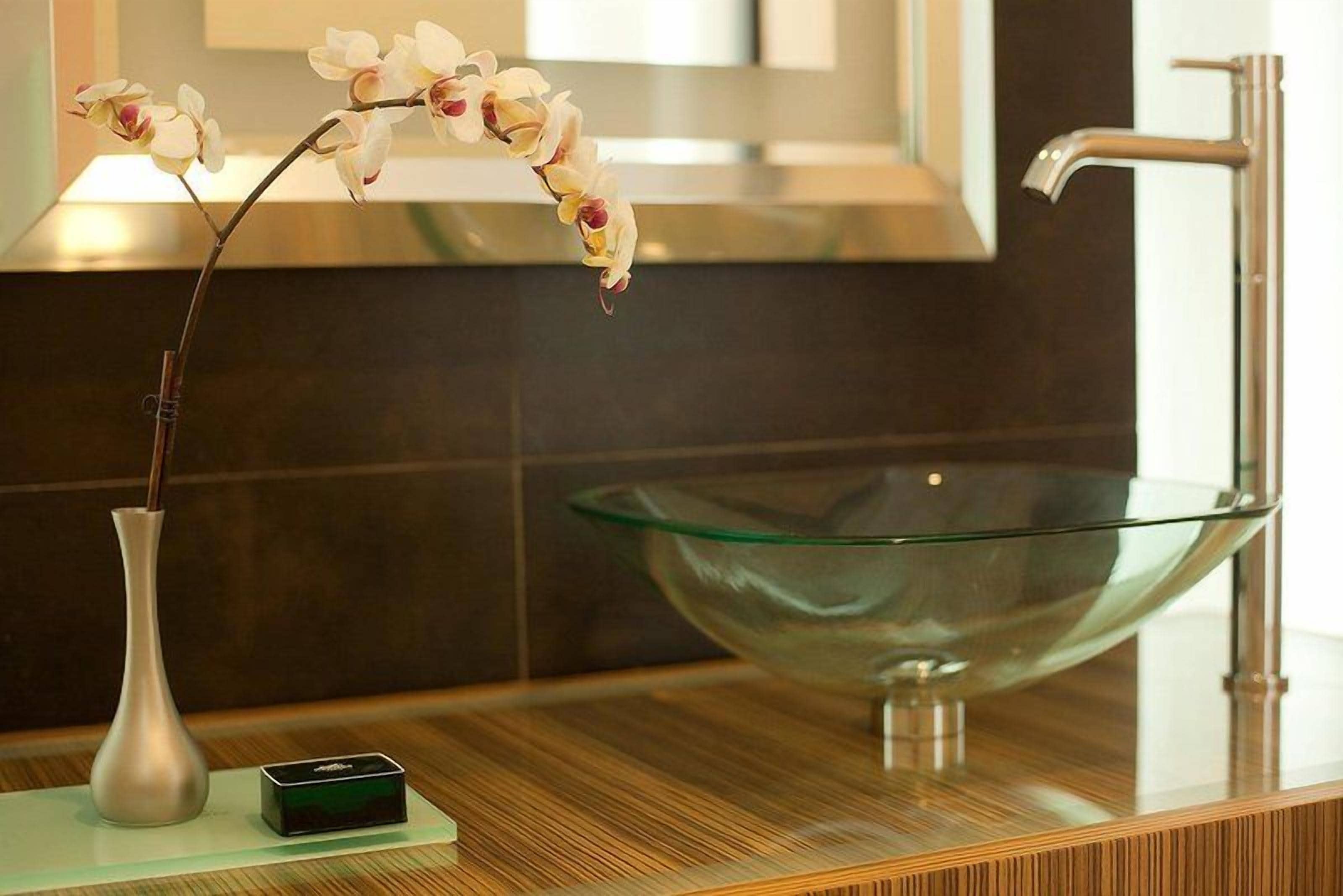 A modern bathroom with a glass bowl sink, a white vase with flowers, and a small electronic device on the counter.