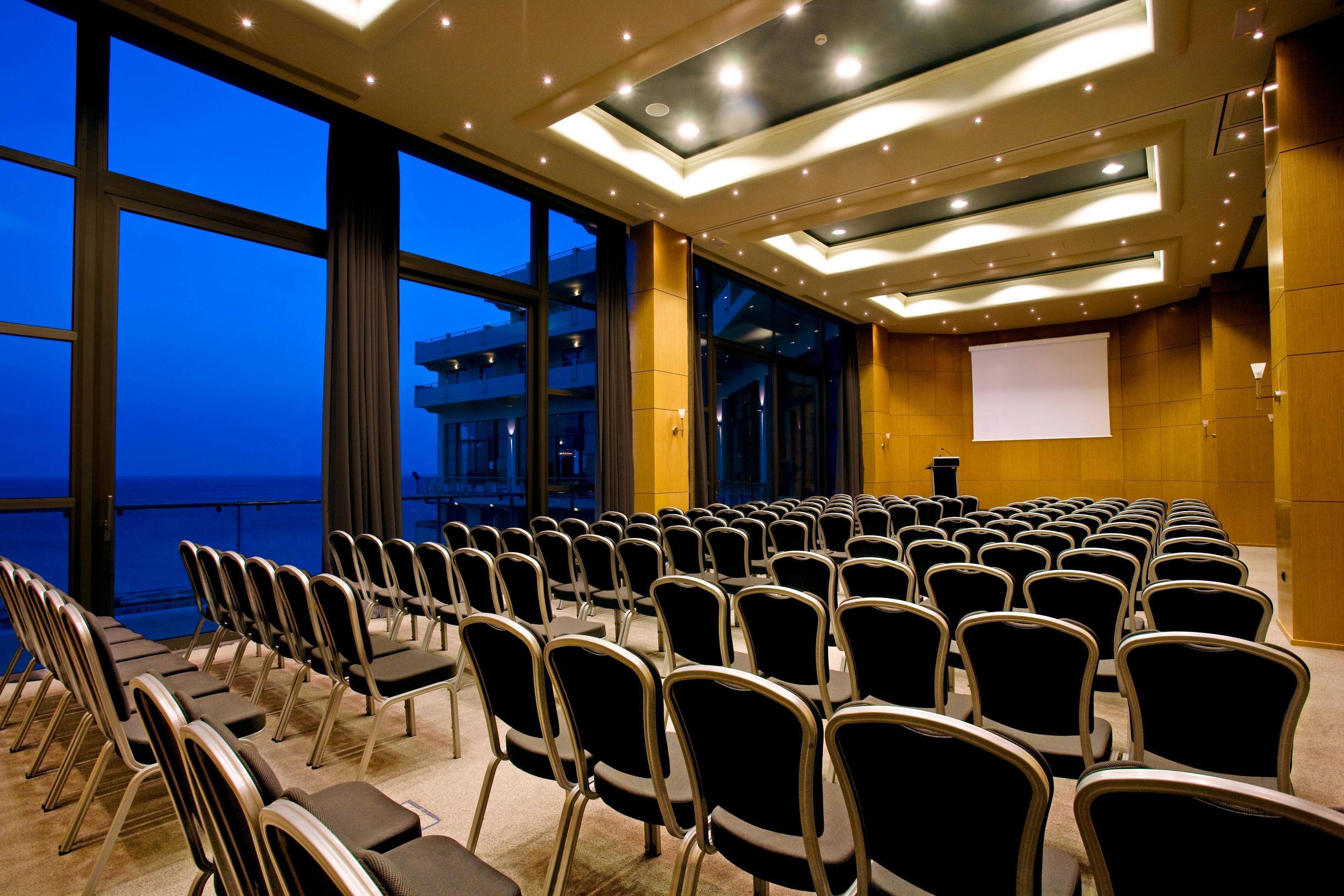 A conference room with rows of chairs facing a projection screen, large windows with a view of the sea, and a modern ceiling design.