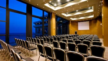 A conference room with rows of chairs facing a projection screen, large windows with a view of the sea, and a modern ceiling design.