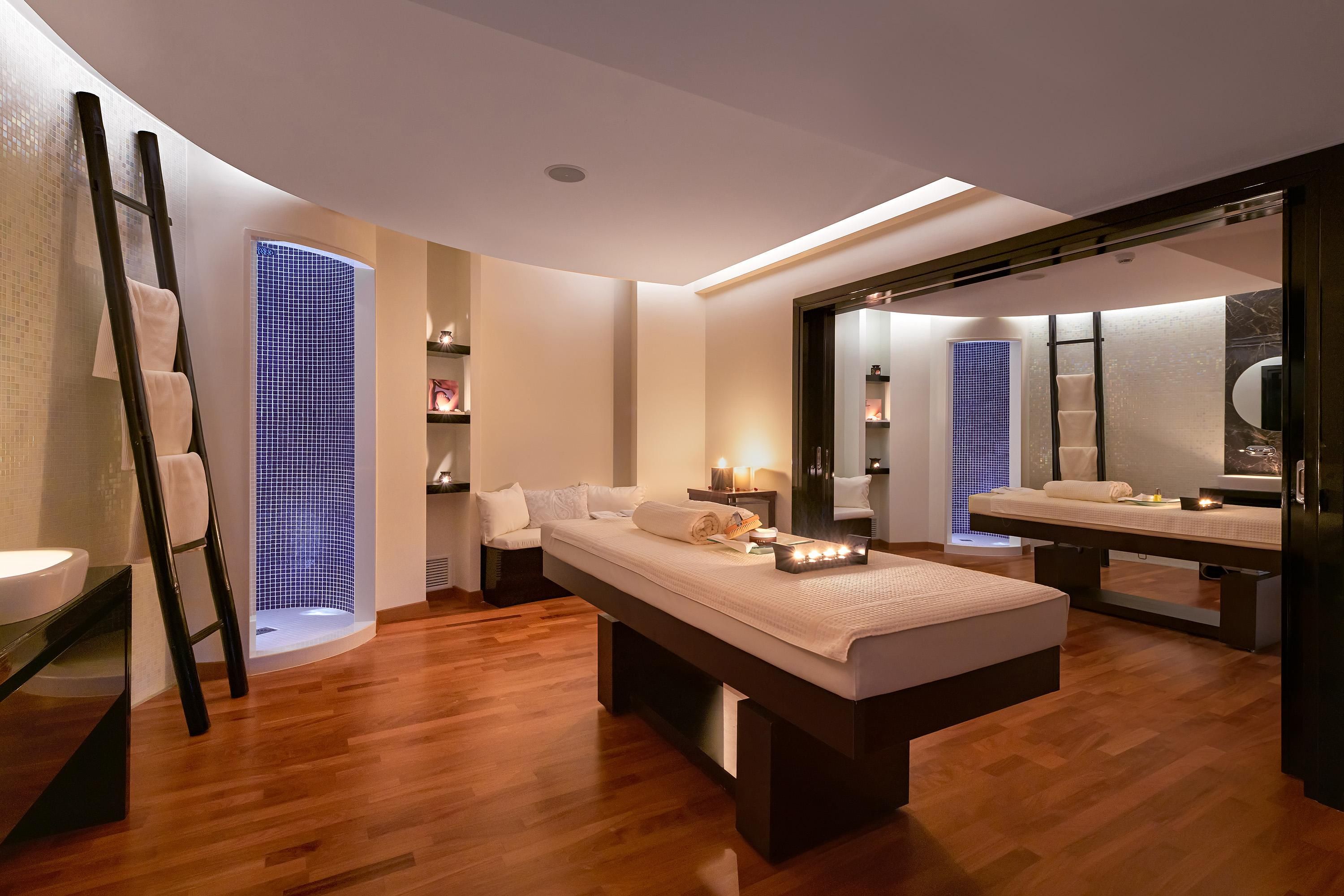 Ein moderner Wellnessraum mit Holzboden, einer Massageliege, einem großen Spiegel und einem Fenster mit blauer Glasbeschichtung.