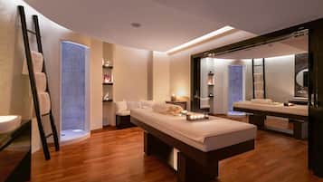 Ein moderner Wellnessraum mit Holzboden, einer Massageliege, einem großen Spiegel und einem Fenster mit blauer Glasbeschichtung.