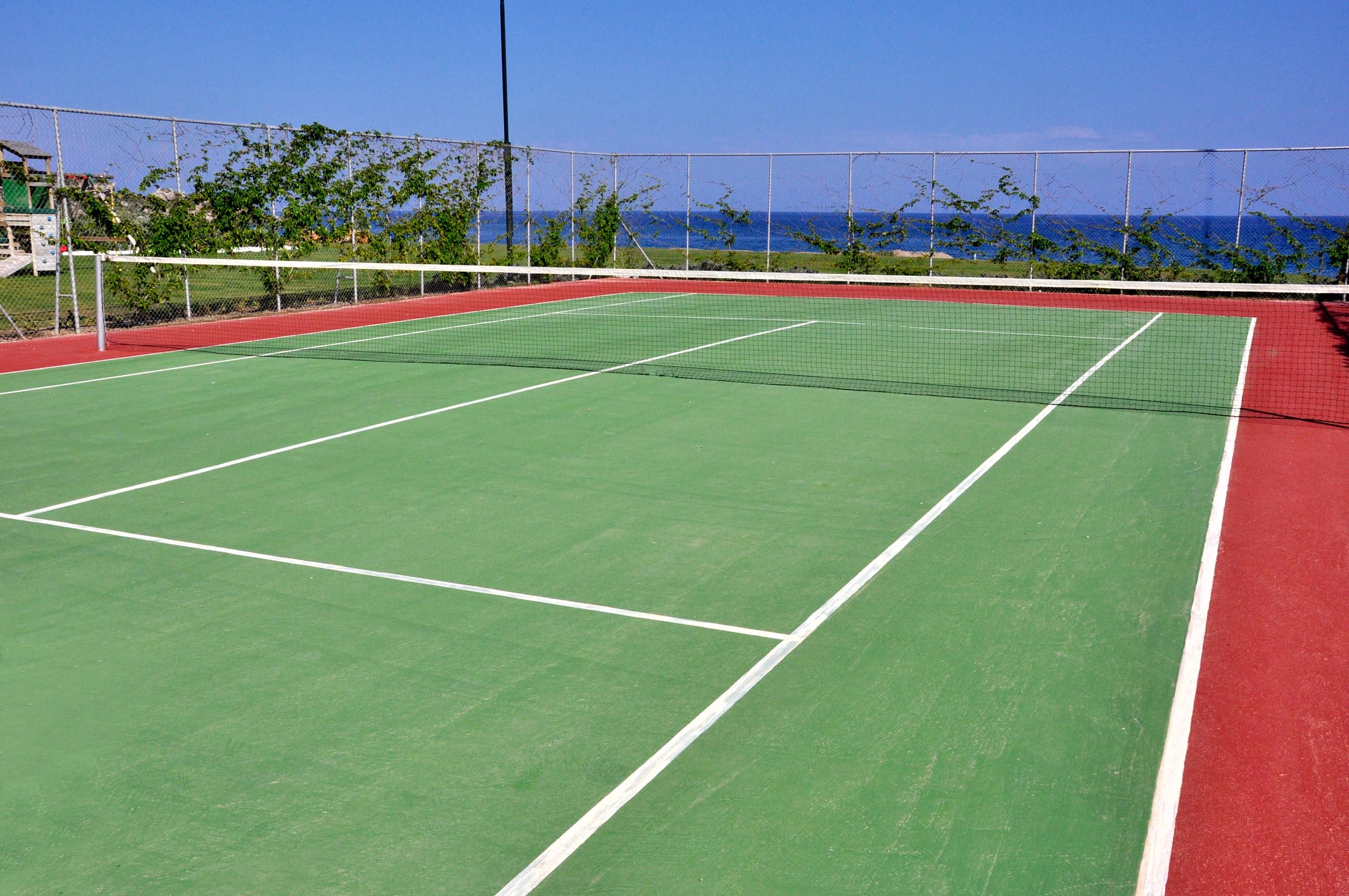 Una cancha de tenis con una red, rodeada por una valla y vista al océano.