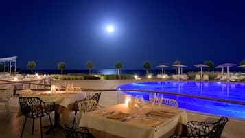 Un área de comedor al aire libre con mesas dispuestas para dos, con vistas a una piscina y al mar bajo un cielo nocturno con una luna visible.
